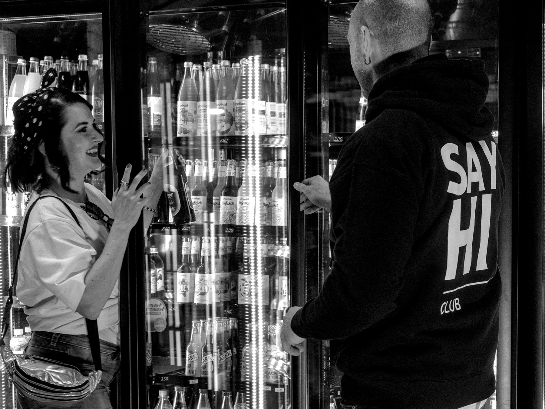 Two people laughing together at a drinks fridge, one wearing a SAY HI Club hoodie